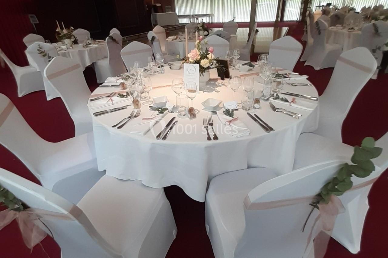 Table ronde élégamment dressée avec nappes blanches, couverts, verres, bougies et centre de table floral.