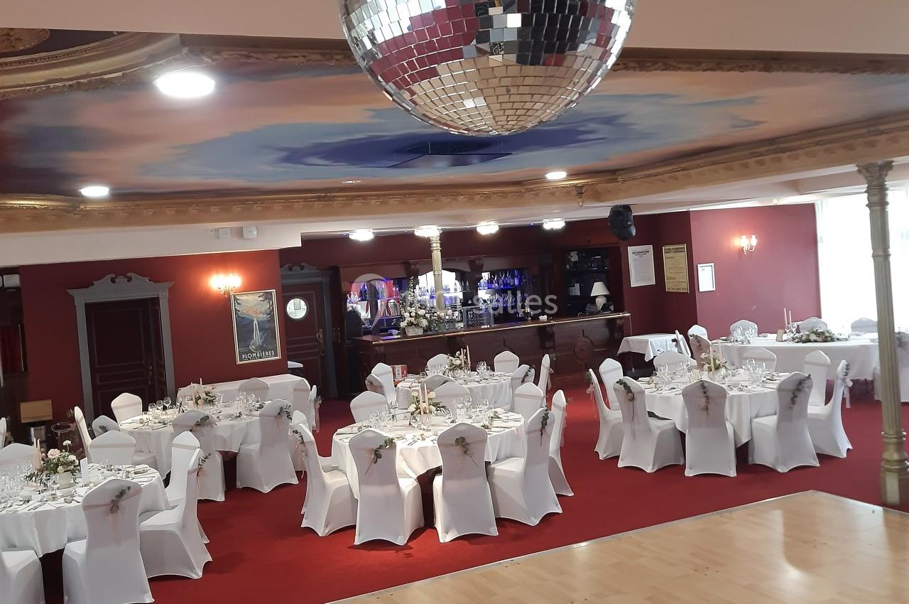 Salle de réception avec tables rondes dressées, chaises blanches et une piste de danse sous une boule à facettes.