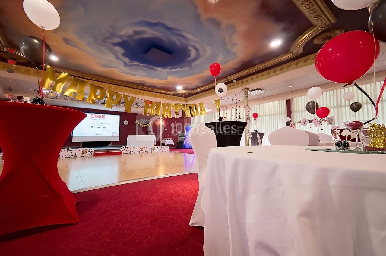 Salle décorée pour une fête, avec ballons rouges et noirs, tables dressées et une bannière ’Happy Birthday’.