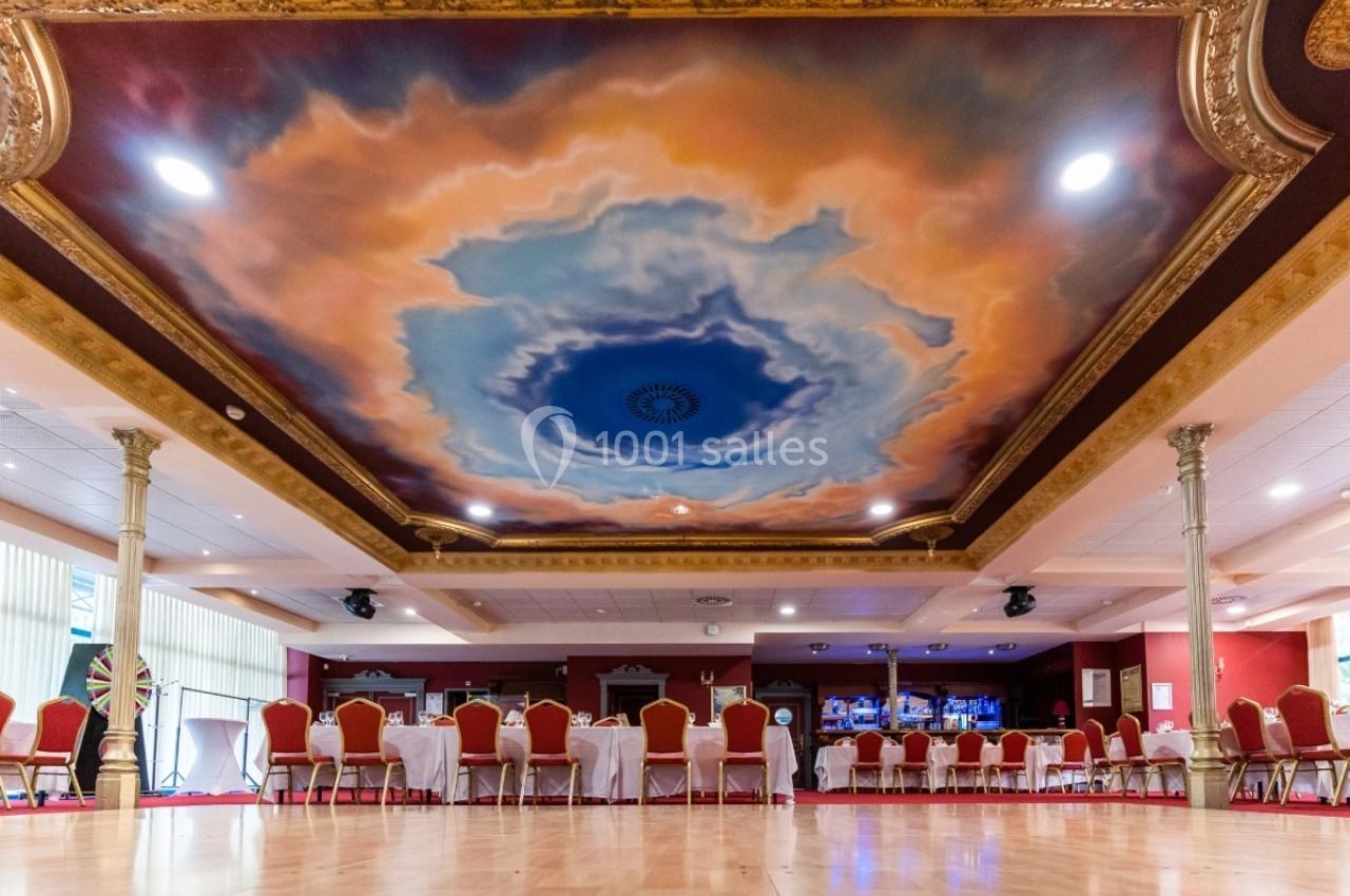 Plafond peint avec un ciel nuageux entouré de moulures dorées, dans une salle ornée de chaises et tables alignées.