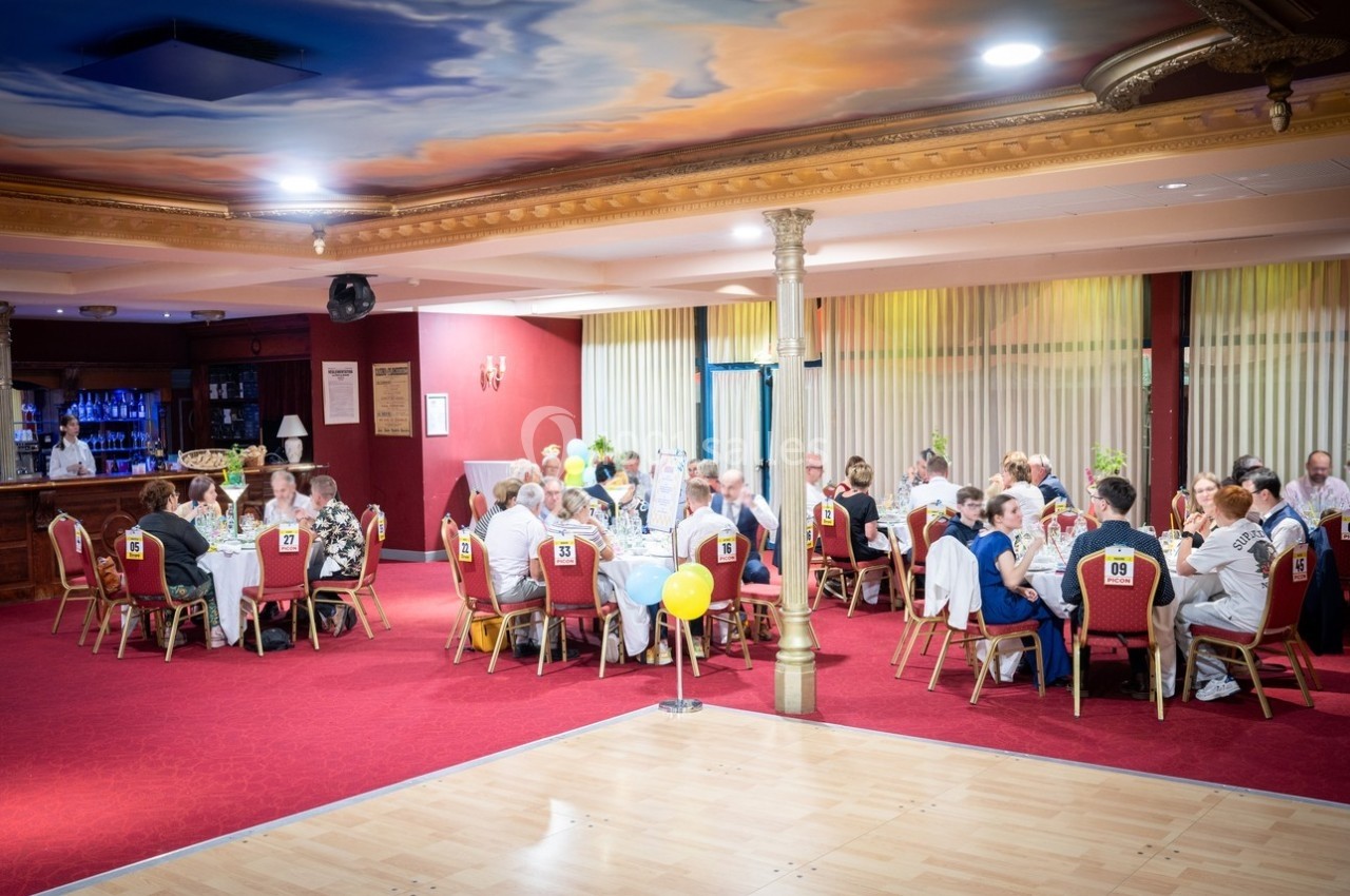 Des convives assis autour de tables rondes dans une salle de réception décorée, avec un plafond peint et un sol en parquet.
