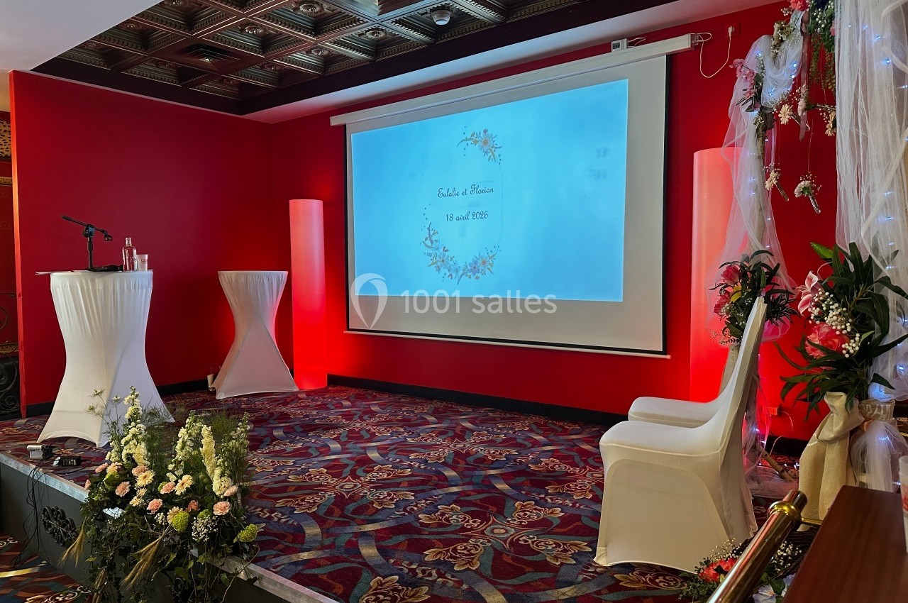 Salle décorée pour un mariage avec écran de projection, fleurs, chaises blanches et éclairage rouge.
