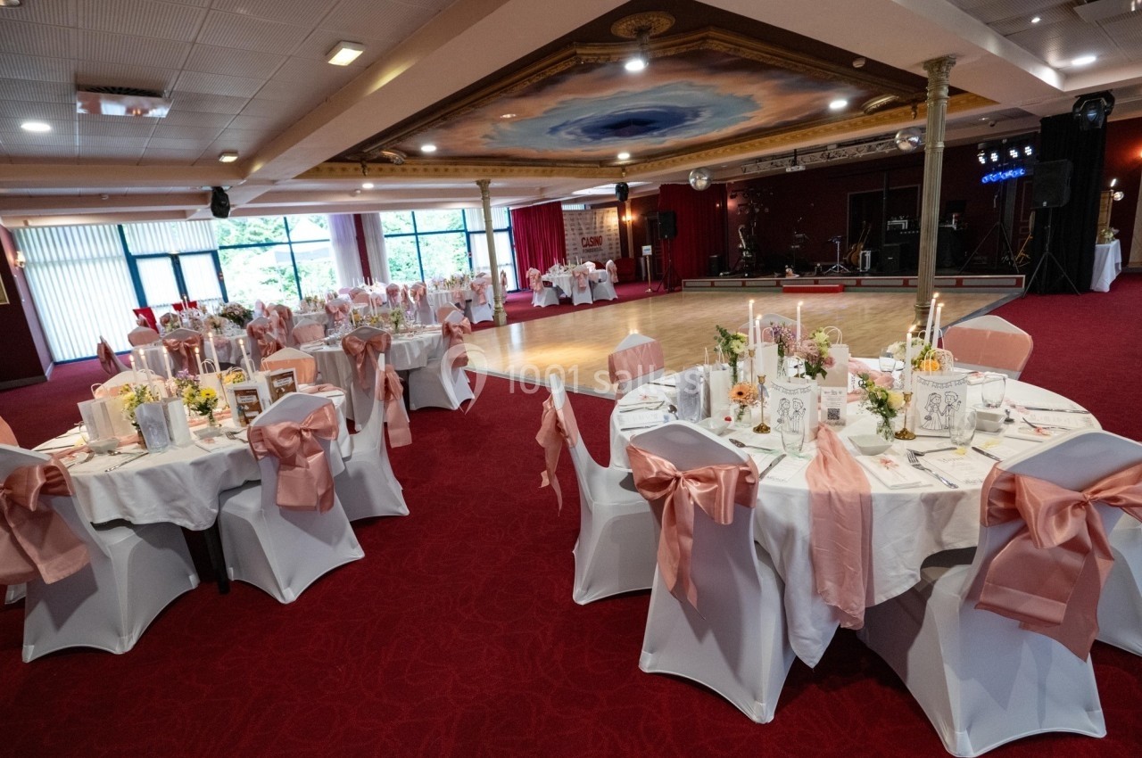 Ambiance rose feutrée Salle de réception décorée avec des tables rondes, nappes blanches, chaises ornées de nœuds roses et bougies allumées.