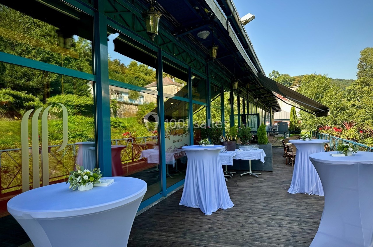 Apéritif sur la terrasse Terrasse en bois avec tables hautes drapées de blanc, bordée de grandes baies vitrées et entourée de verdure.
