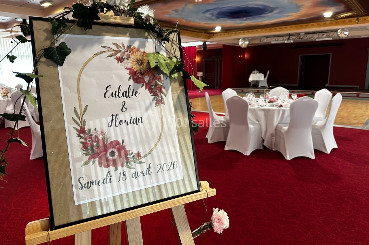 Bienvenue à votre mariage Panneau décoratif annonçant un mariage avec des fleurs, posé sur un chevalet dans une salle de réception.