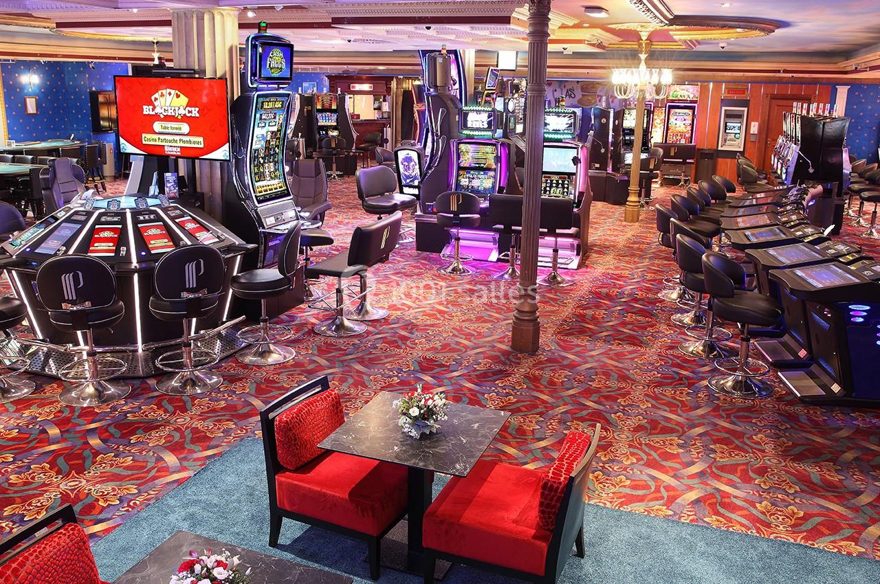 Salle de casino avec machines à sous, tables de jeux électroniques et sièges rouges sur un sol à motifs colorés.