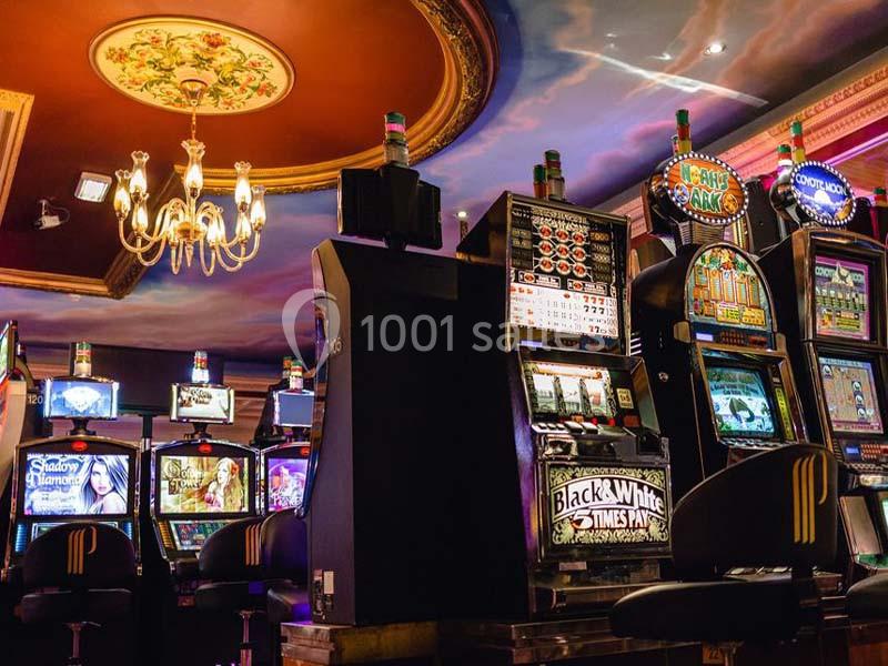 Machines à sous, côté jeux Salle de jeux avec des machines à sous alignées sous un plafond décoré et un lustre suspendu.