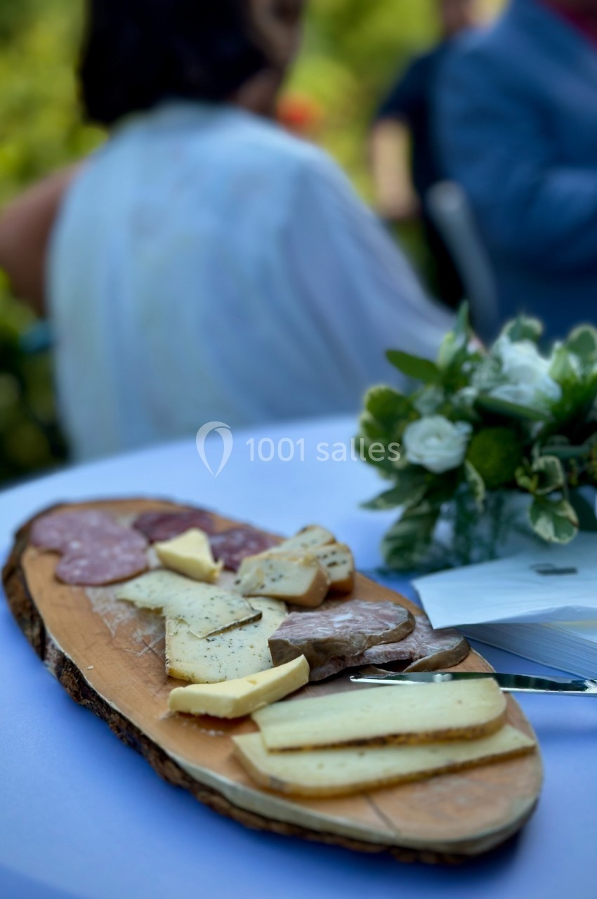 Moment agréable à partager en terrasse Plateau de charcuterie et de fromages variés sur une planche en bois, posé sur une table blanche avec un bouquet de fleurs.