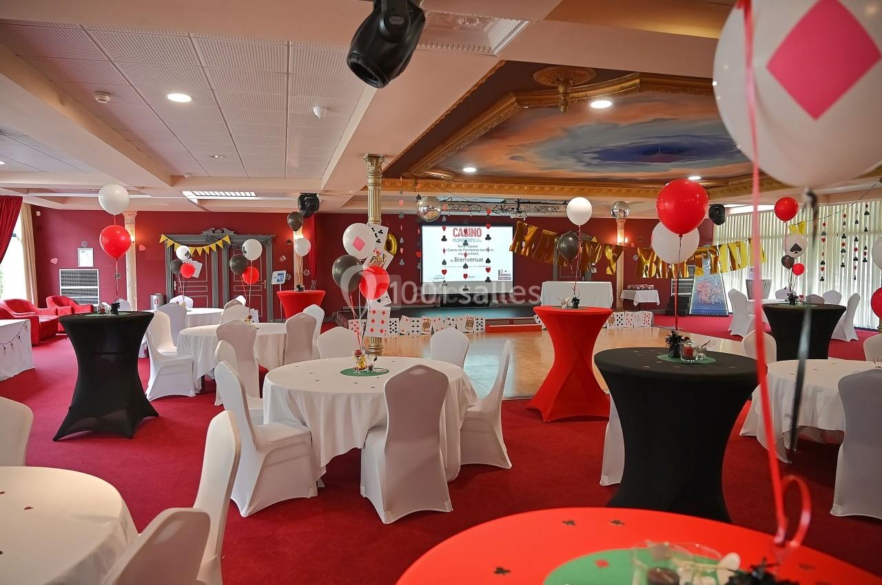 Salle décorée sur le thème du casino avec tables rondes, nappes blanches, ballons et écran de projection.