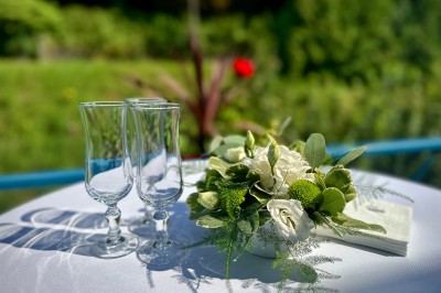 Miniature Sur la terrasse surplombant la rivière Deux verres à pied et un bouquet de fleurs blanches et vertes sur une table ronde en extérieur.