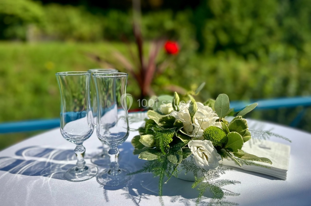 Sur la terrasse surplombant la rivière Deux verres à pied et un bouquet de fleurs blanches et vertes sur une table ronde en extérieur.