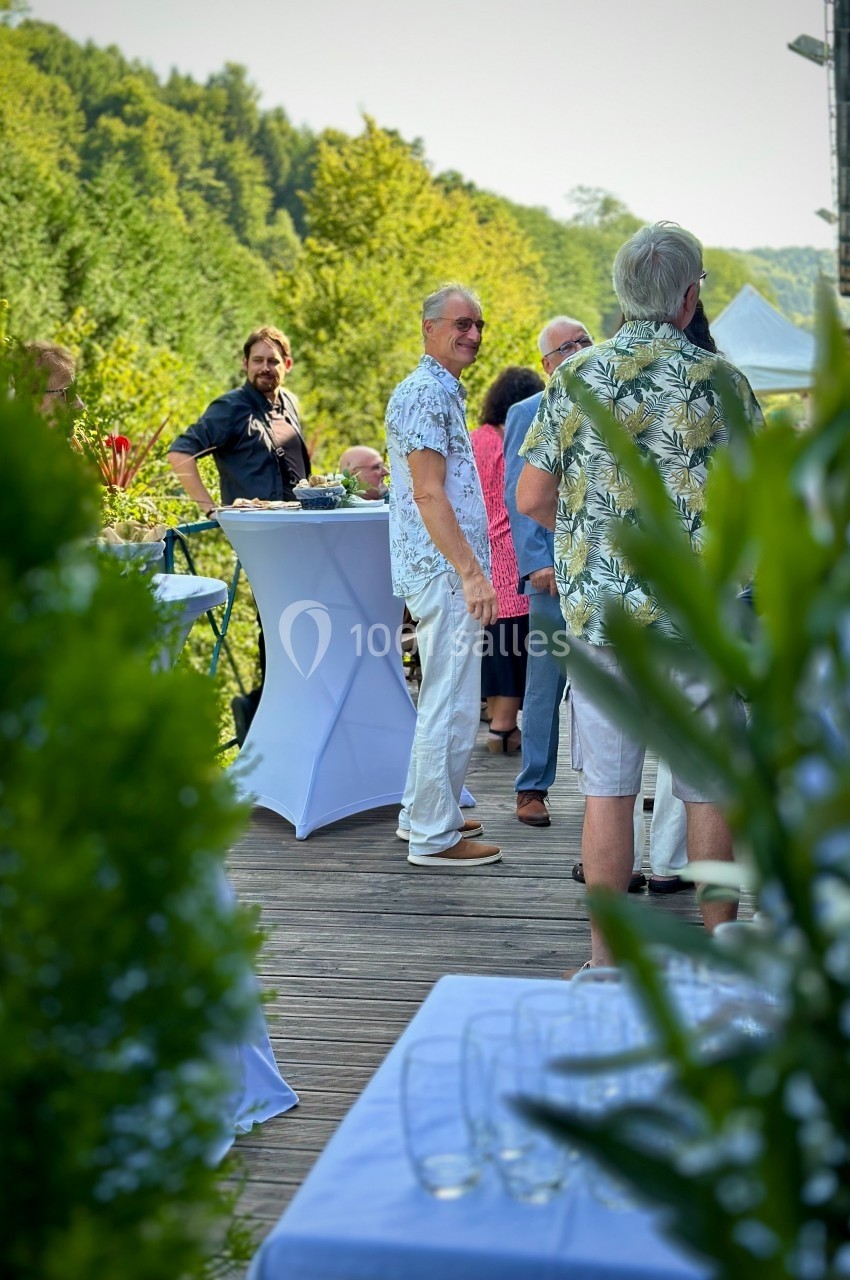 Vin d'honneur en terrasse Des personnes discutent debout autour de tables hautes sur une terrasse en bois entourée de verdure.