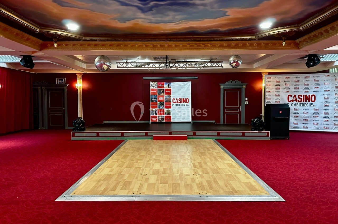 Salle avec une petite scène, piste de danse en bois, moquette rouge et plafond décoré de nuages peints.