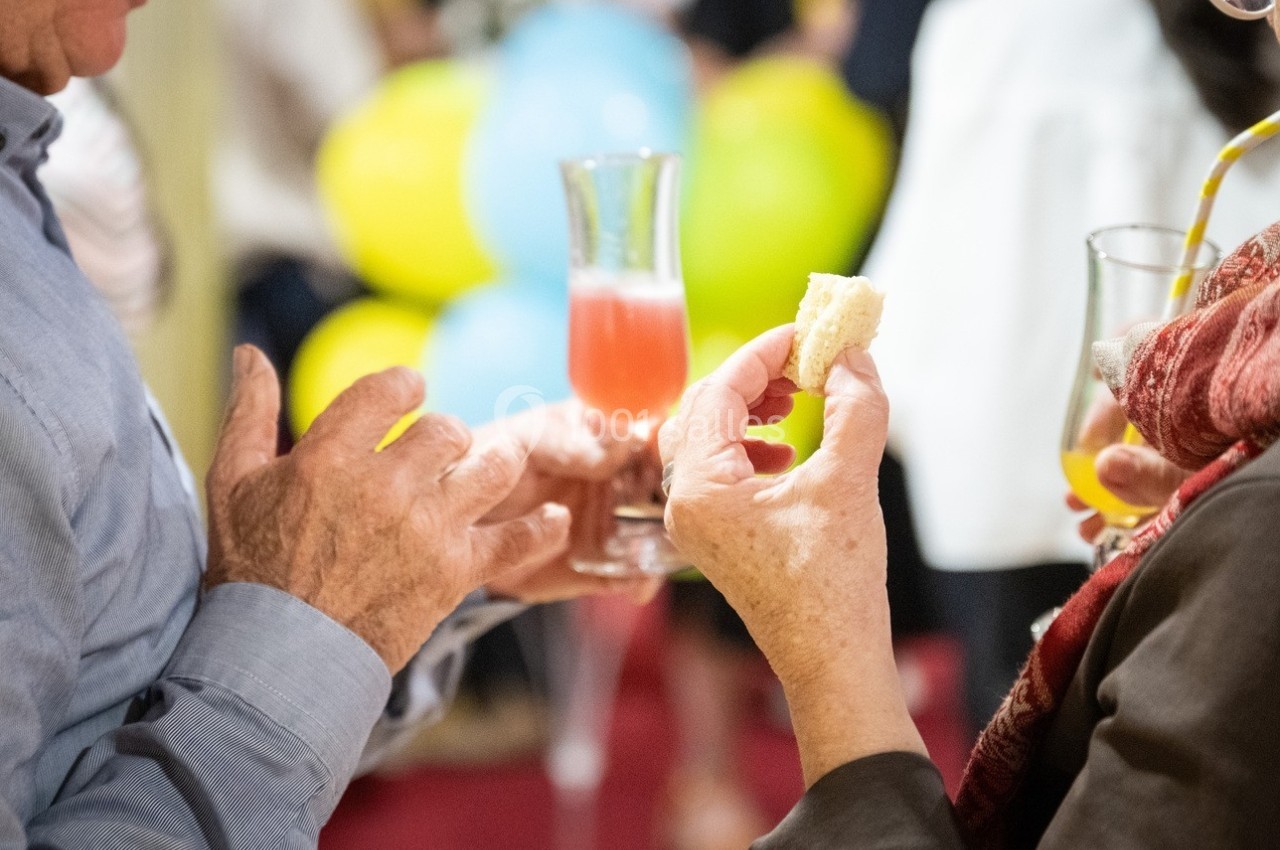 Deux personnes discutent en tenant des verres et un morceau de pain lors d'un événement avec des ballons en arrière-plan.