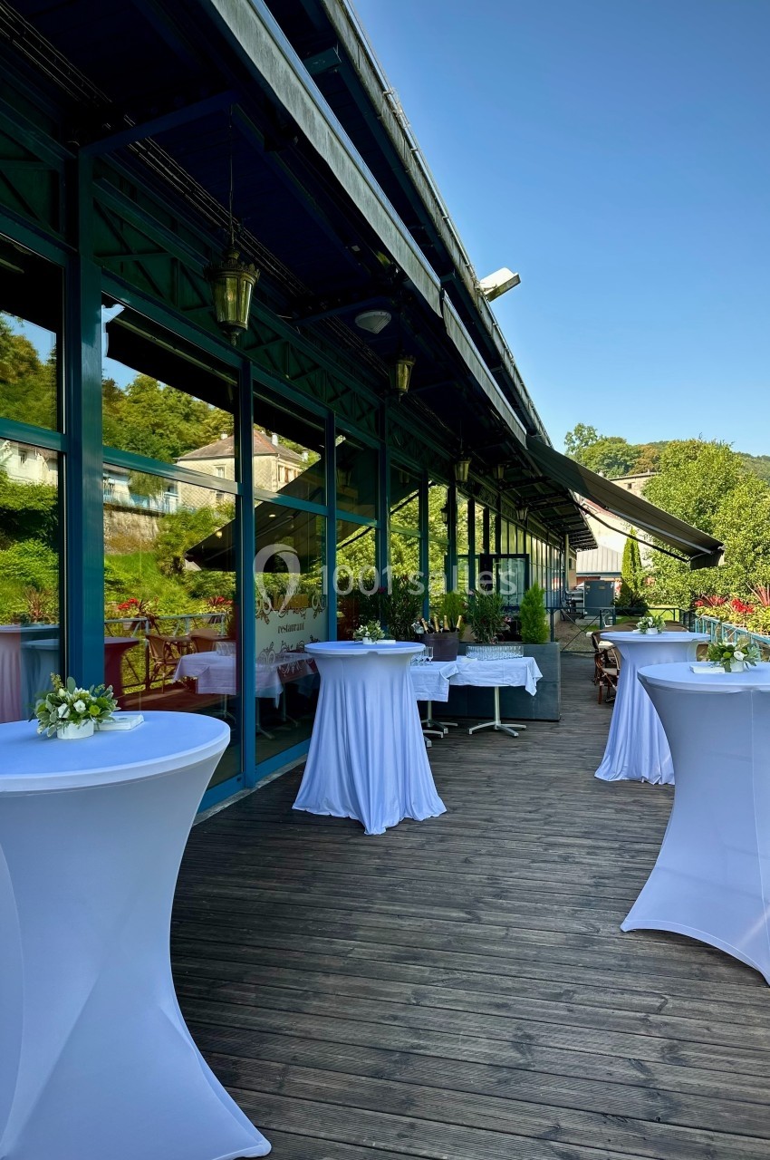 Terrasse en bois avec tables hautes drapées de blanc, entourée de verdure et jouxtant un bâtiment vitré.
