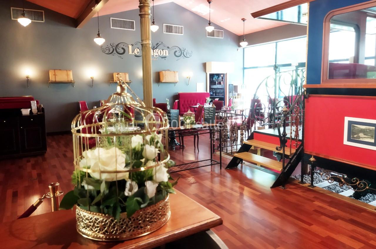 Salle de restaurant élégante avec parquet, mobilier rouge, escalier en fer forgé et fleurs blanches en cage dorée.