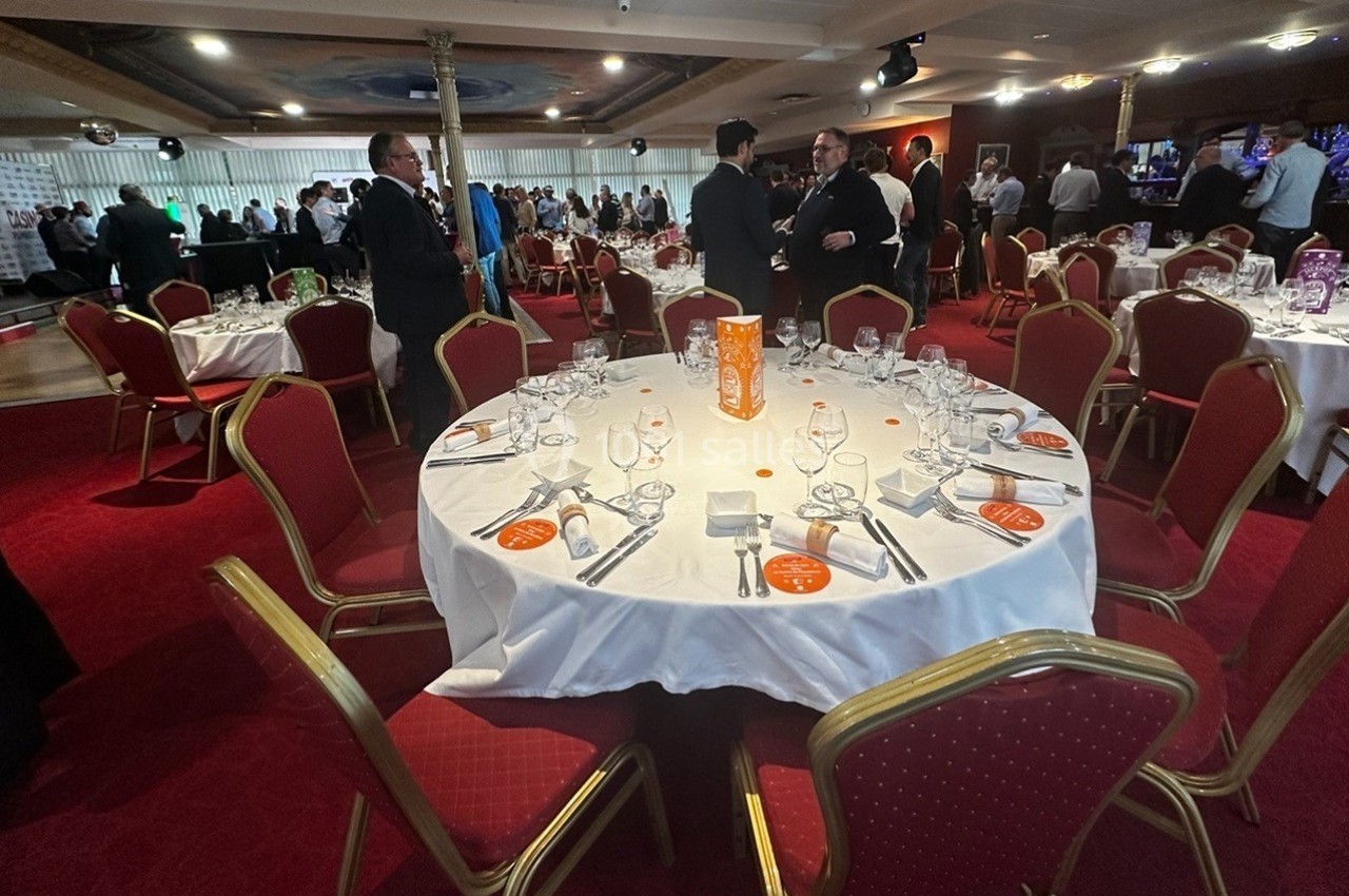 Dîner de gala Salle de réception avec tables rondes dressées, nappes blanches, chaises rouges et invités debout en arrière-plan.
