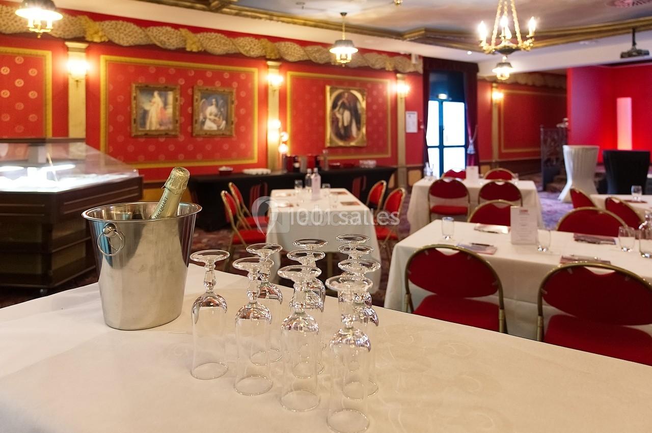 Salle de réception avec tables dressées, chaises rouges, seau à champagne et verres alignés sur une nappe blanche.