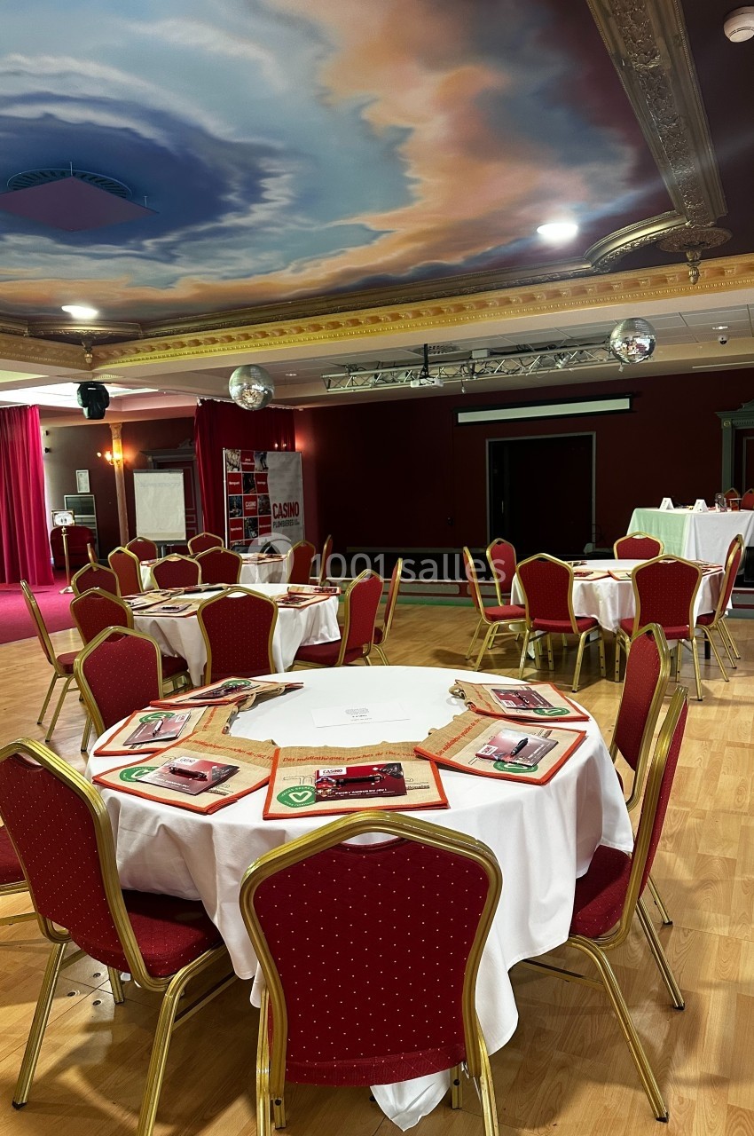 En mode journée d'étude ou réunion Salle de réception avec tables rondes dressées, nappes blanches, chaises rouges et plafond peint en ciel nuageux.