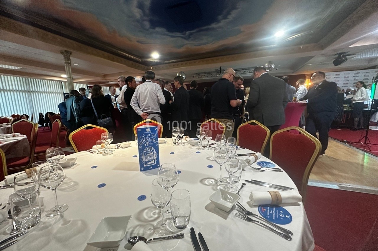 Esprit festif avant un repas servi à table Salle de réception avec des tables dressées au premier plan et un groupe de personnes debout en discussion à l'arrière-plan.