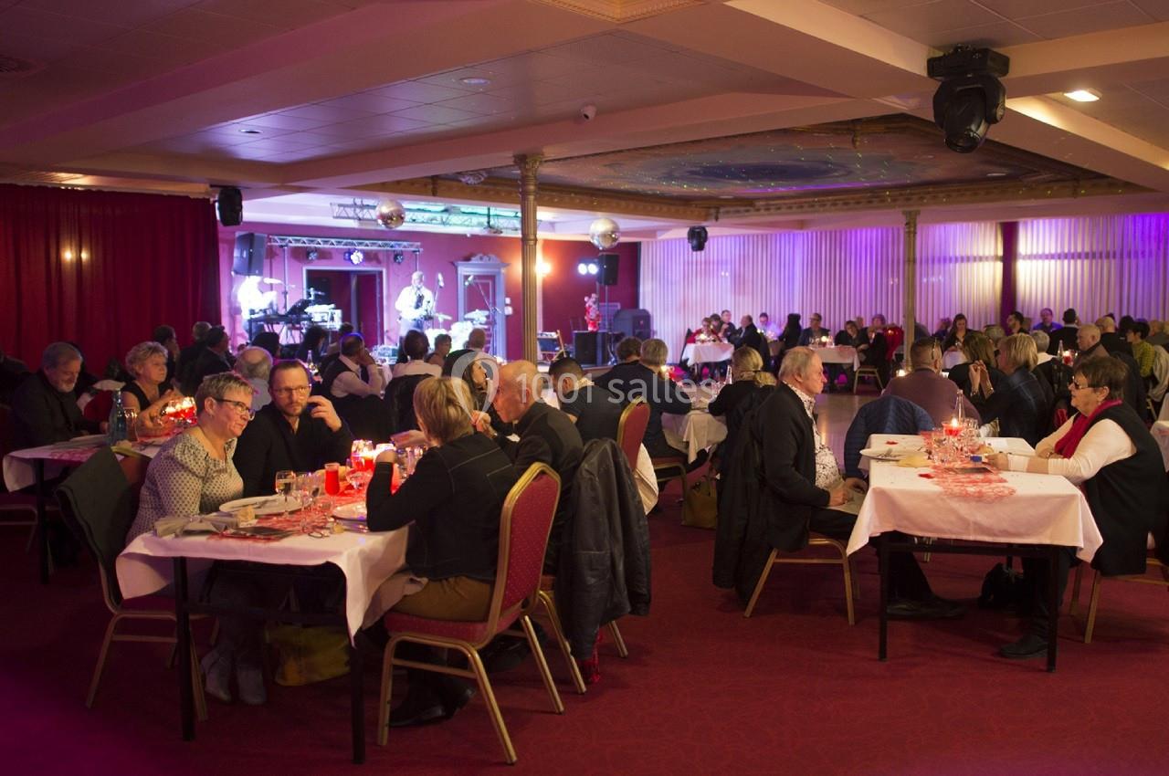 Salle de réception avec des convives assis à des tables, éclairée par des lumières tamisées et une scène en arrière-plan.