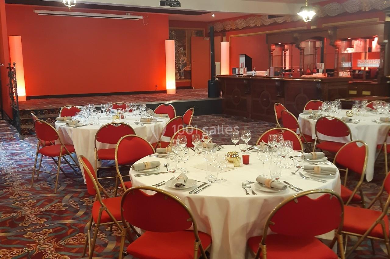 Salle de réception avec tables rondes dressées, chaises rouges et éclairage chaleureux.