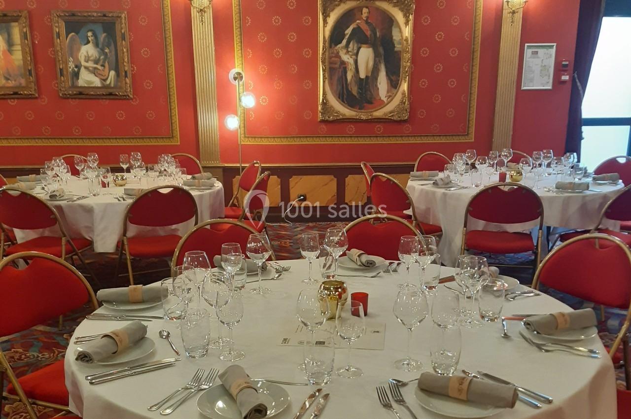 Salle de réception élégante avec tables rondes dressées, nappes blanches, chaises rouges et décorations murales classiques.