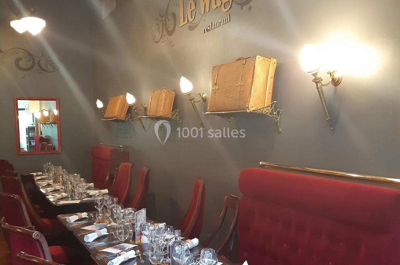 Salle de restaurant avec murs gris, banquettes rouges, tables dressées et valises décoratives sur des étagères murales.