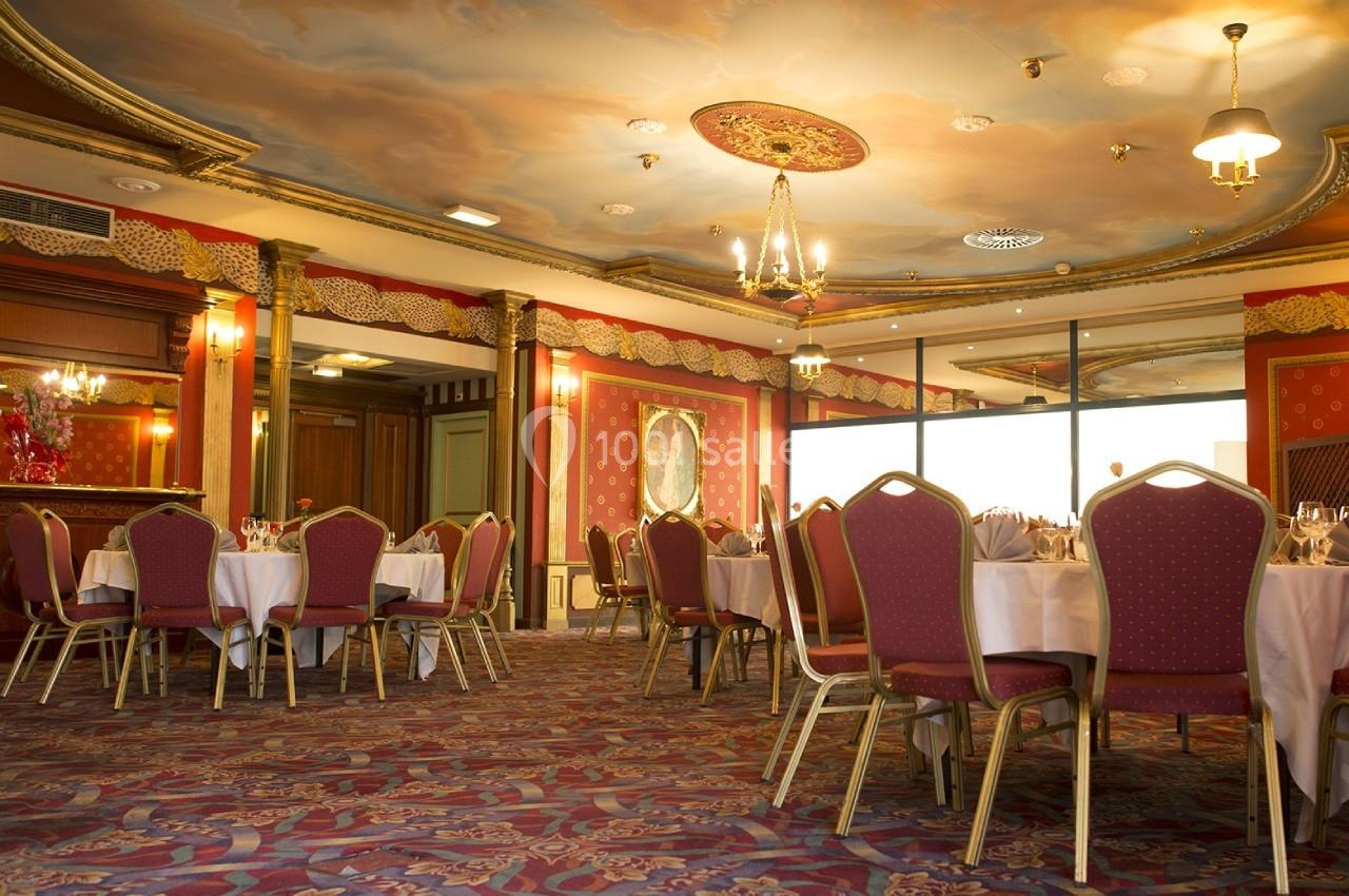 Salle de réception élégante avec tables rondes dressées, chaises rouges et plafond décoré de fresques.