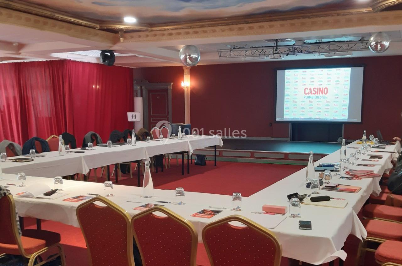 Salle de réunion avec tables disposées en U, chaises rouges, écran de projection et rideaux rouges en arrière-plan.