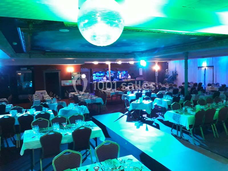 Salle de réception éclairée avec des tables dressées, une piste centrale et un bar illuminé en arrière-plan.