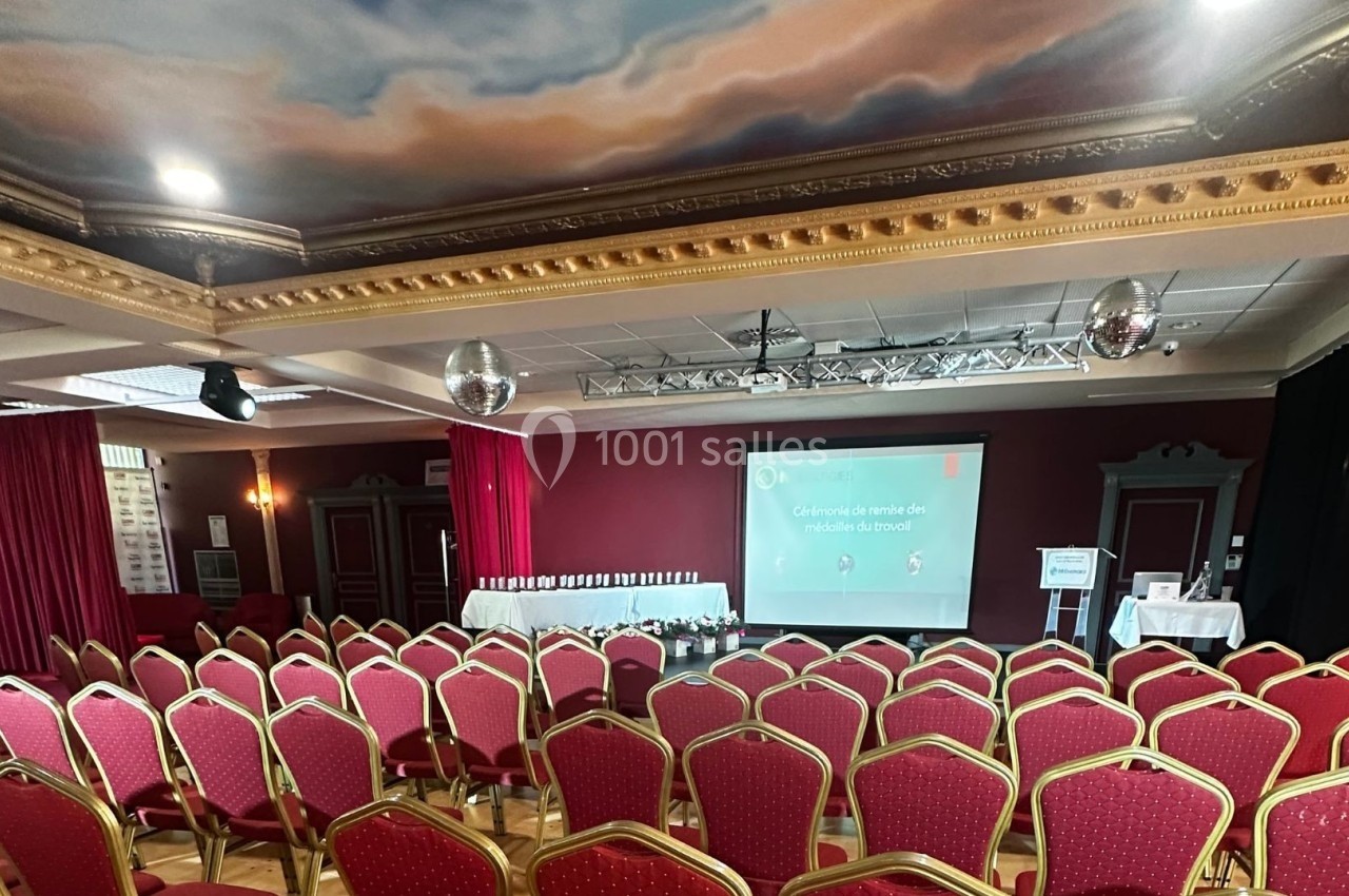 Mise à disposition scène, éclairage, sono et micros Salle de conférence avec rangées de chaises rouges, écran de projection allumé et décorations au plafond.