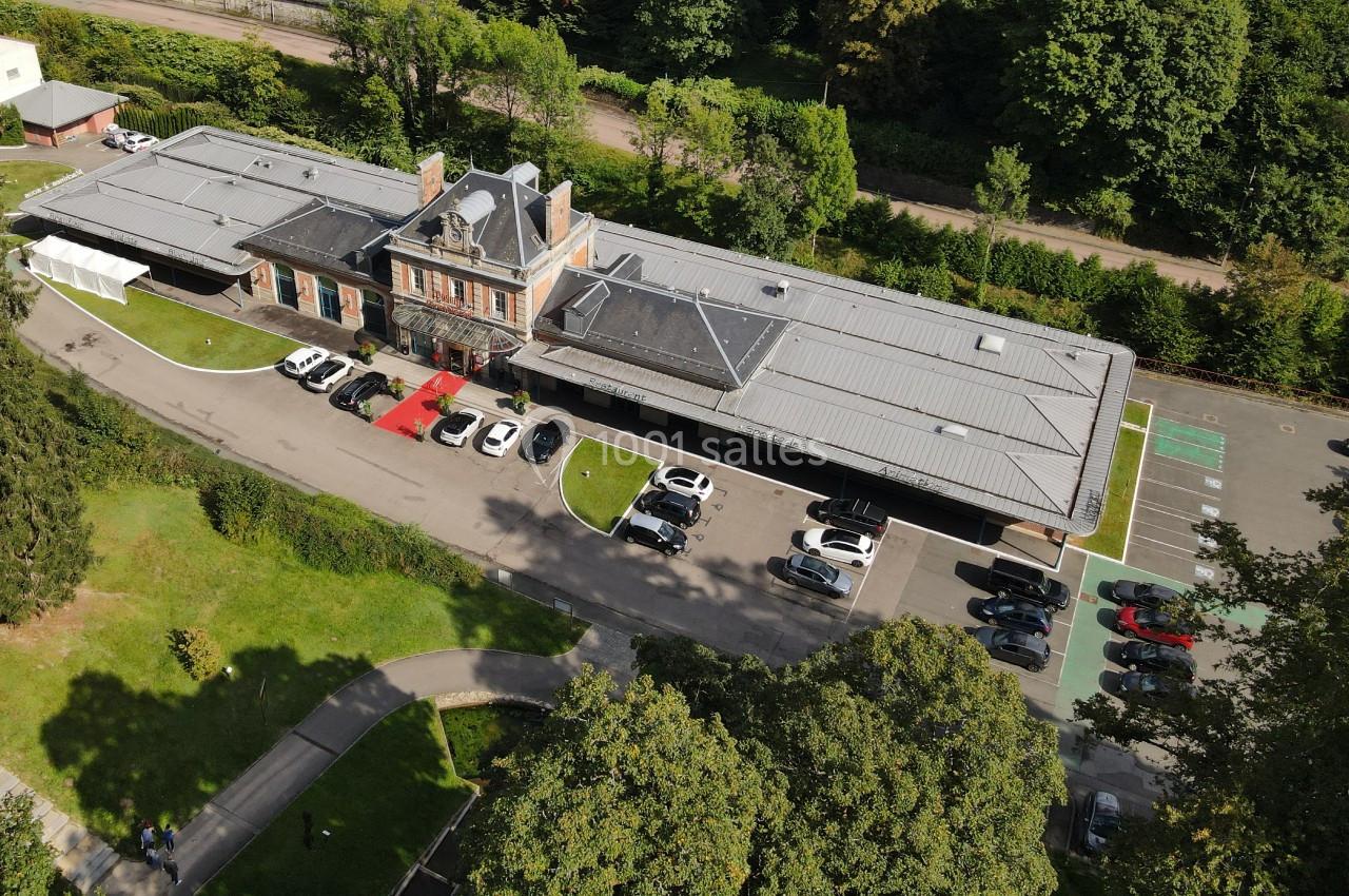 Vue aérienne d'un bâtiment en briques avec un toit en métal, entouré de voitures garées et de verdure.