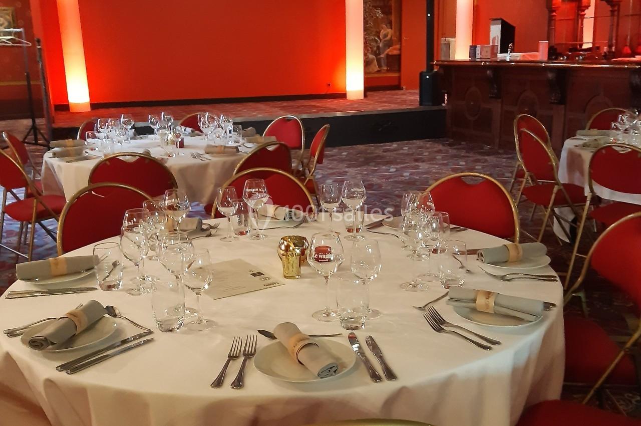 Salle de réception avec tables rondes dressées, nappes blanches, chaises rouges et éclairage mural orange.