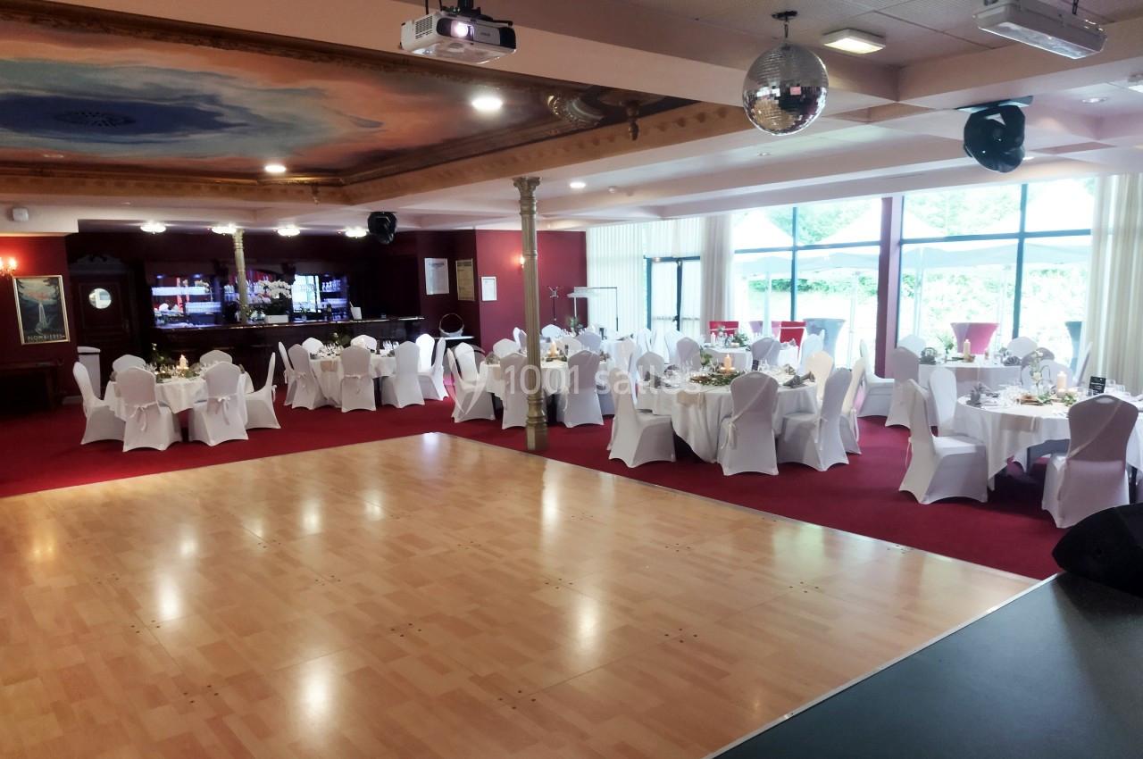 Salle de réception avec tables dressées, chaises blanches, piste de danse en bois et bar en arrière-plan.