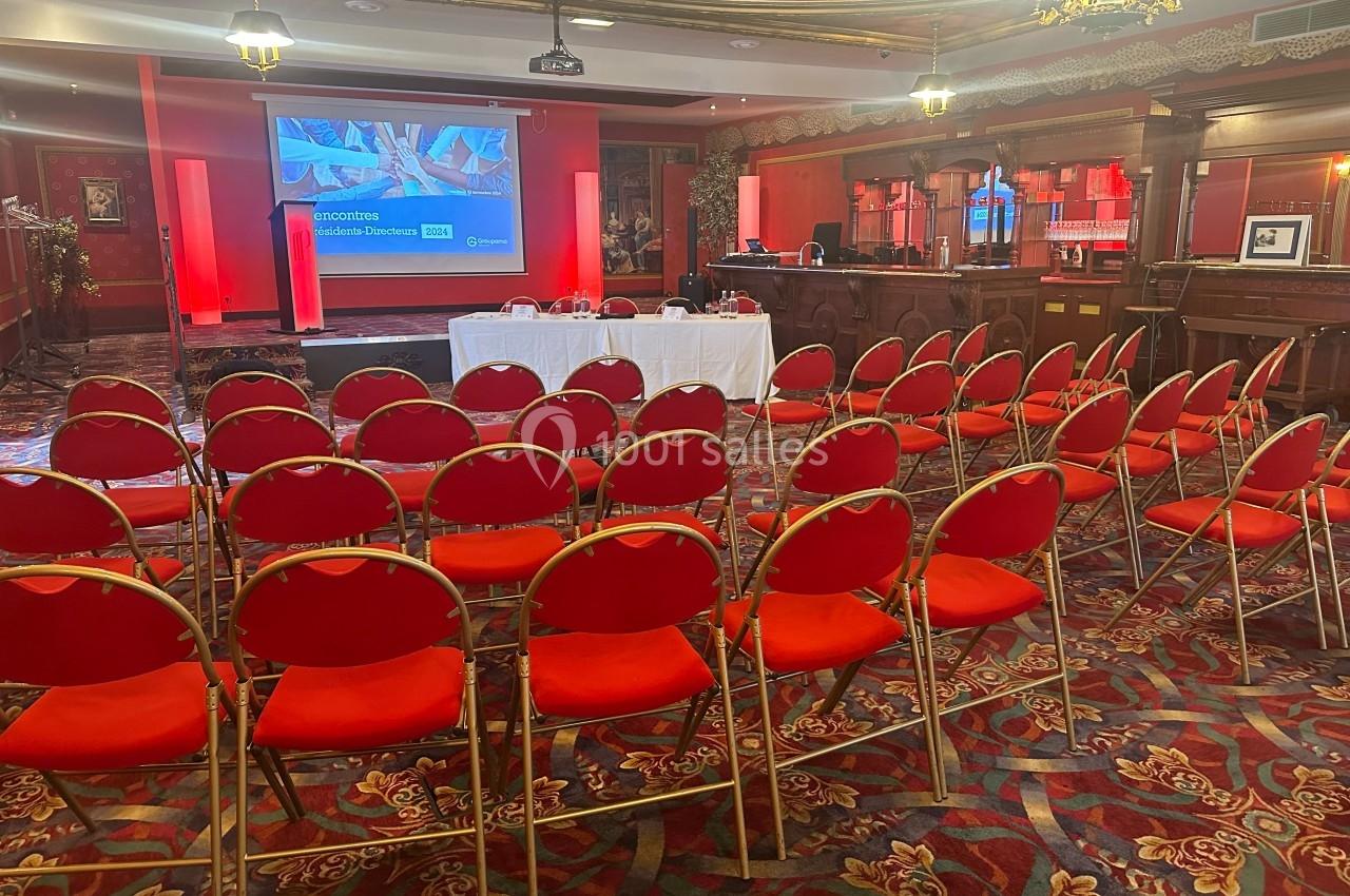Salle de conférence avec rangées de chaises rouges, écran de présentation allumé et table blanche à l'avant.