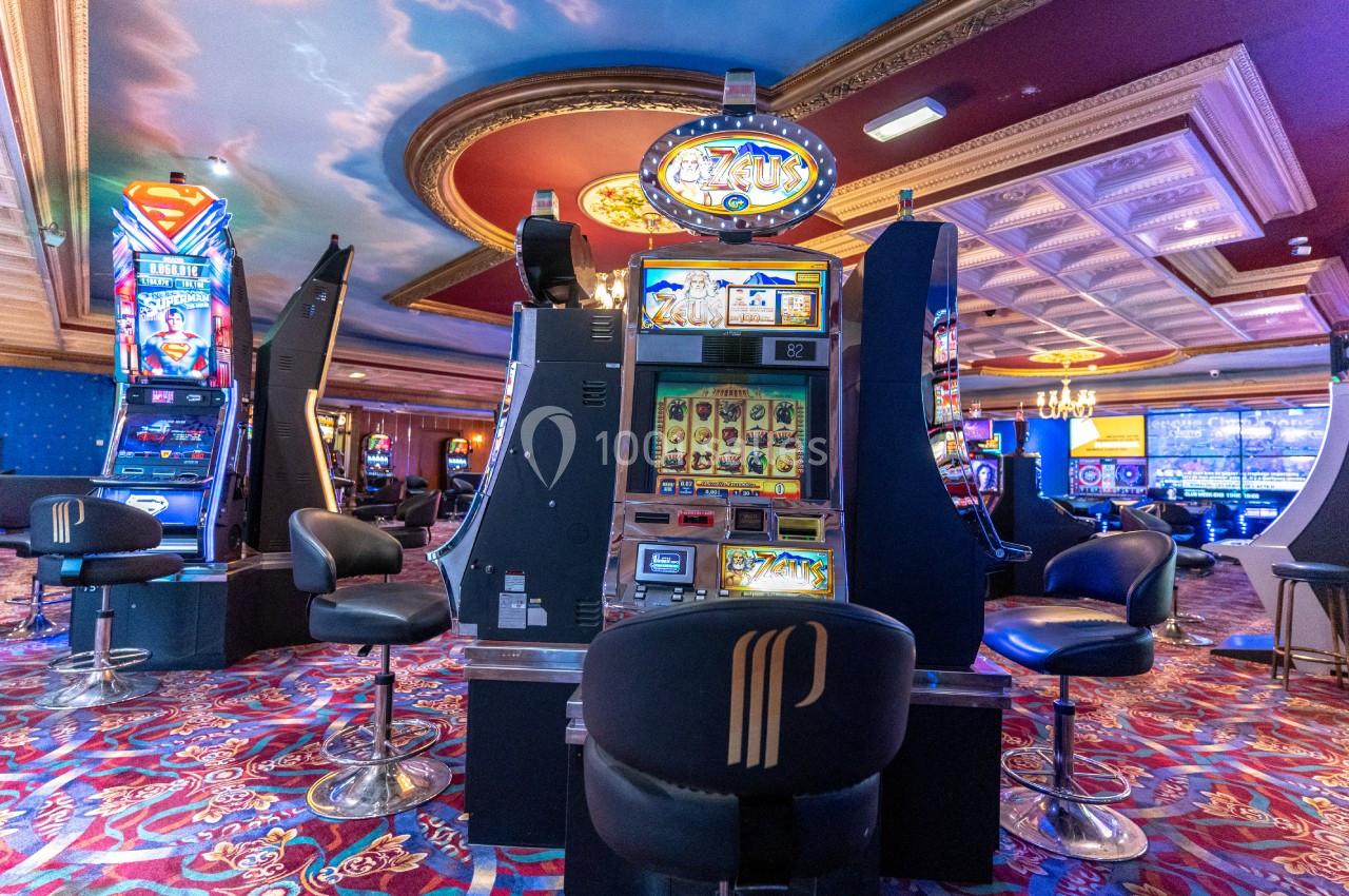 Salle de casino avec machines à sous illuminées, sièges rembourrés et plafond décoré de fresques.