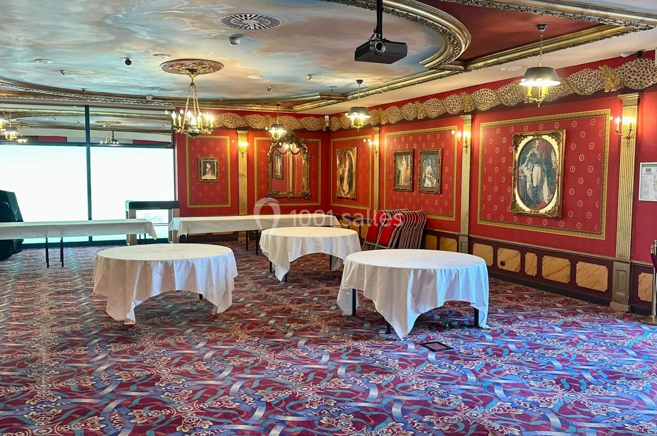 Salle décorée de rouge avec tapis à motifs, tables recouvertes de nappes blanches et cadres accrochés aux murs.