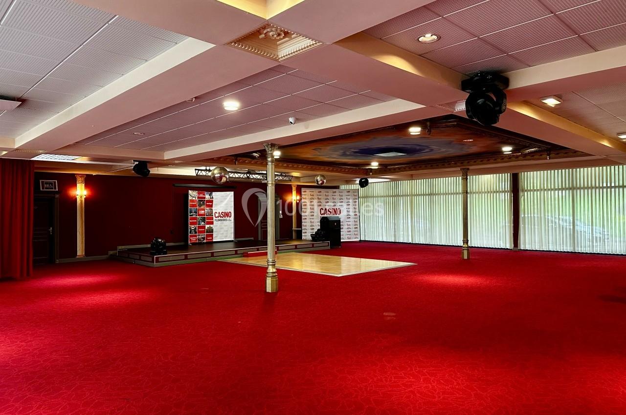 Salle spacieuse avec moquette rouge, scène surélevée, rideaux et grandes baies vitrées laissant entrer la lumière naturelle.