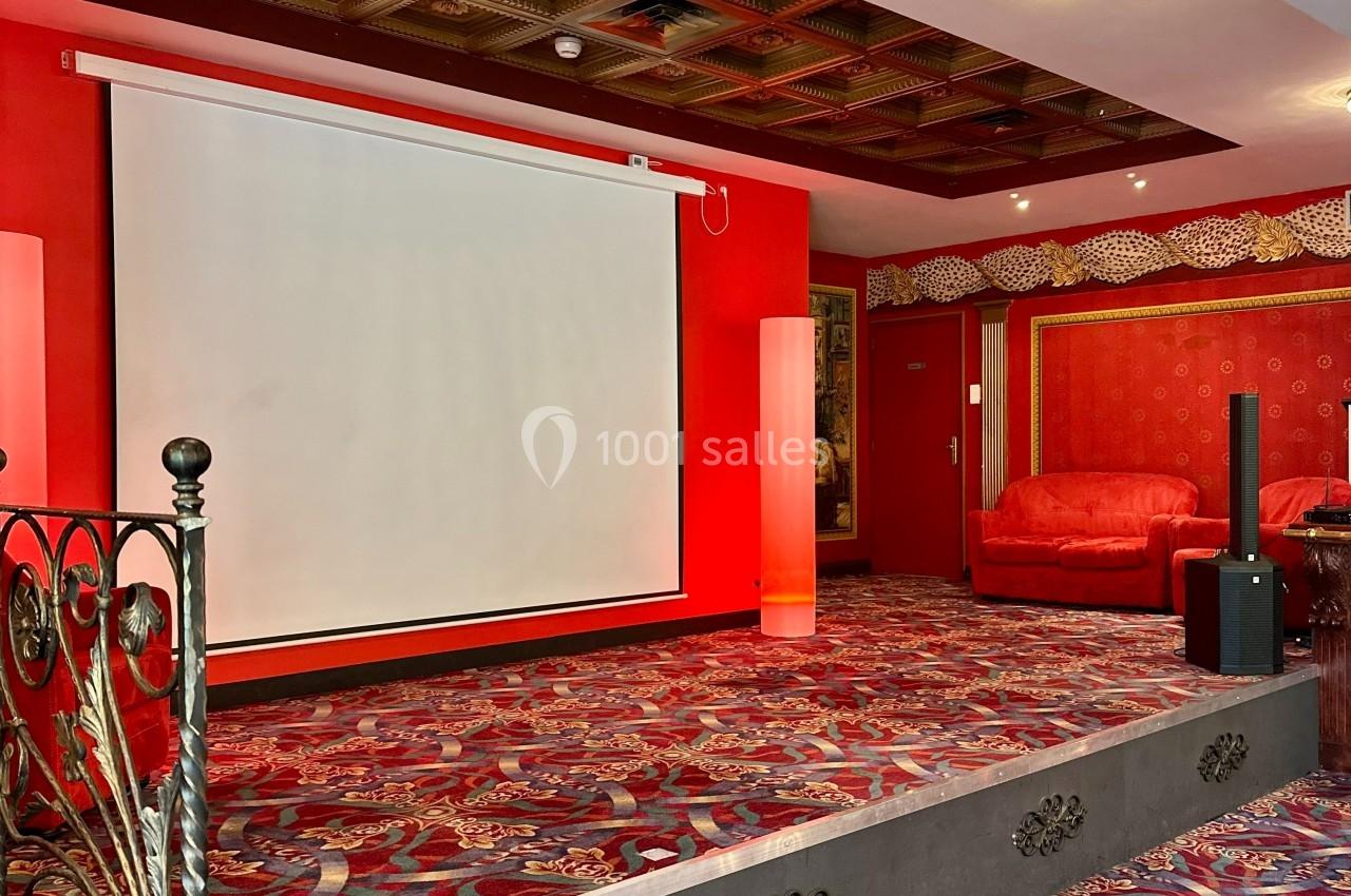 Salle avec écran de projection, tapis rouge à motifs, fauteuils assortis et plafond à caissons décoratifs.