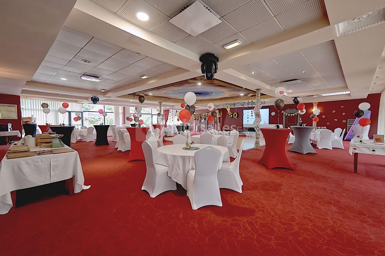 Salle décorée pour un événement avec tables rondes, nappes blanches, ballons colorés et éclairage chaleureux.