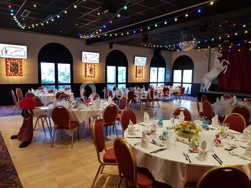 Salle de réception décorée avec des tables dressées, des guirlandes lumineuses et une scène avec un cheval blanc.