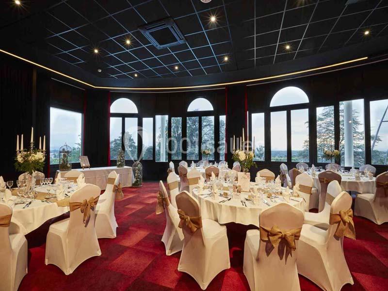Salle de réception élégante avec tables rondes décorées, nappes blanches et grandes baies vitrées donnant sur l'extérieur.