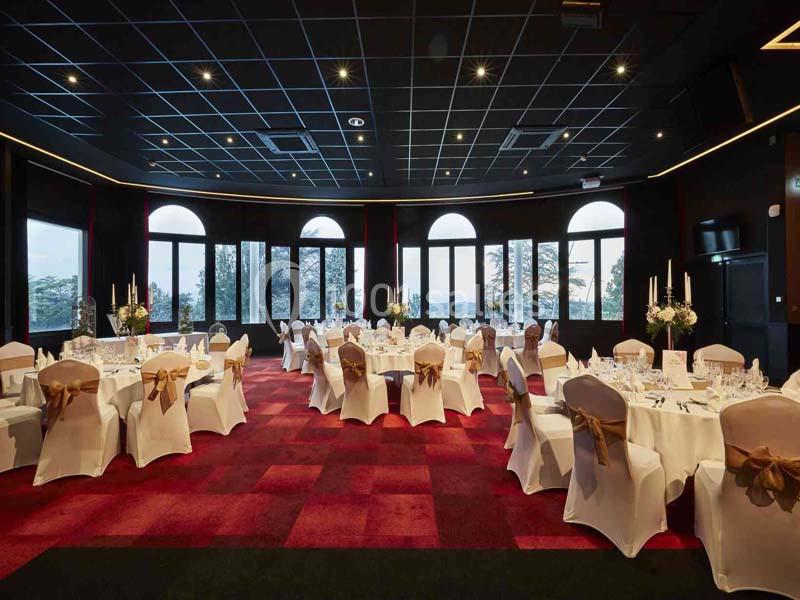 Salle de réception élégante avec tables rondes dressées, nappes blanches et grandes fenêtres donnant sur un paysage.