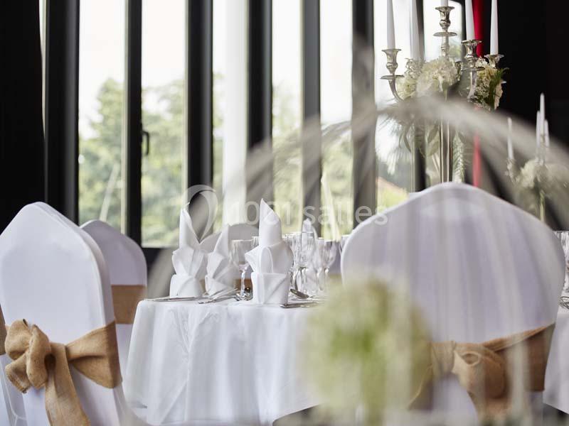 Salle de réception avec tables rondes dressées, nappes blanches, serviettes pliées et chaises décorées de rubans.