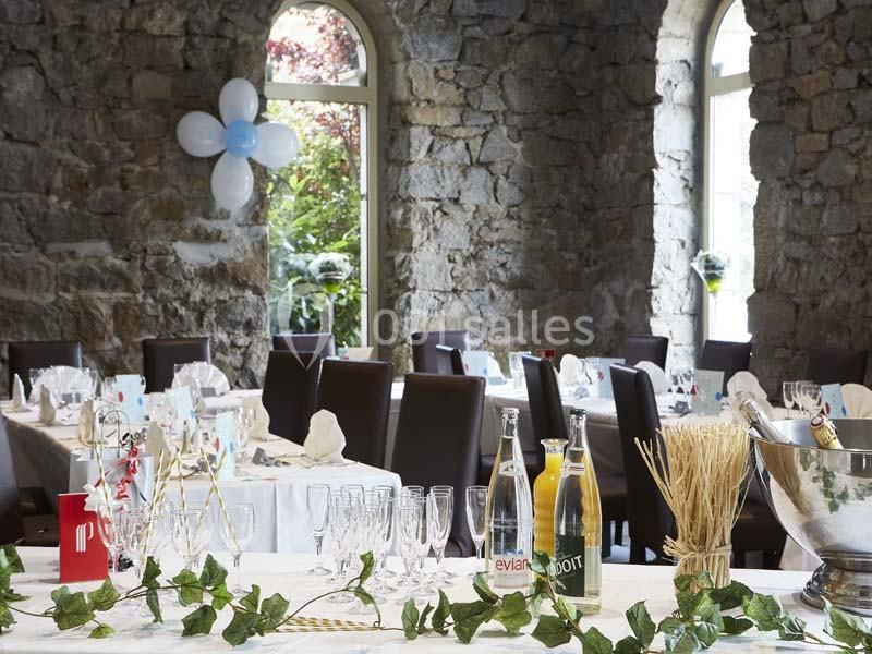 Salle en pierre avec tables dressées pour un événement, décorée de ballons et de feuillage, lumière naturelle entrant par…