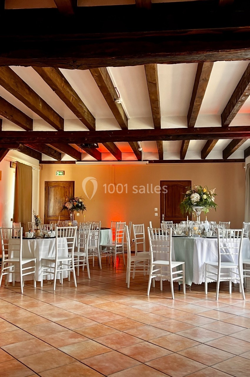 Salle de réception avec tables rondes dressées, chaises blanches et poutres apparentes au plafond.