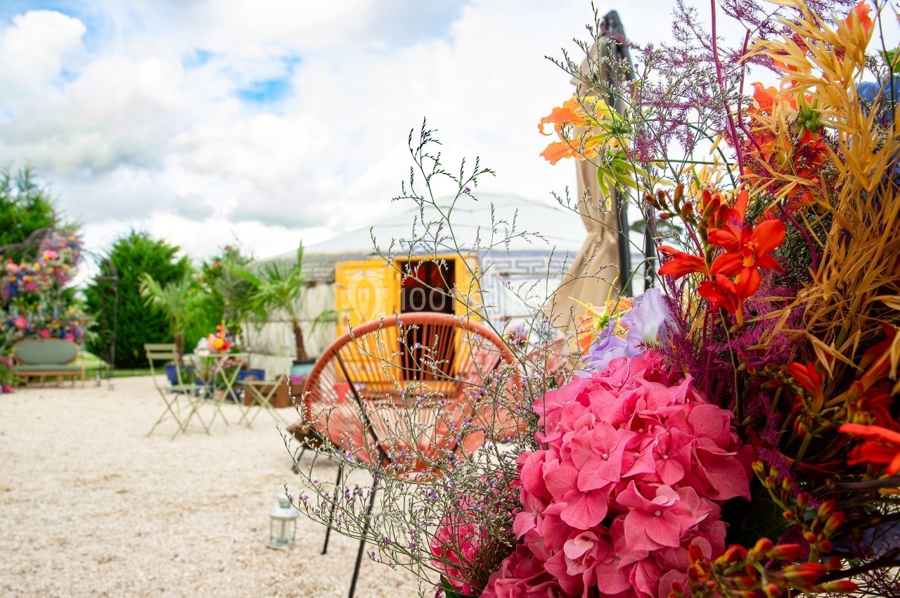 Fleurs colorées au premier plan avec une terrasse extérieure, des chaises et une yourte en arrière-plan sous un ciel nuageux.