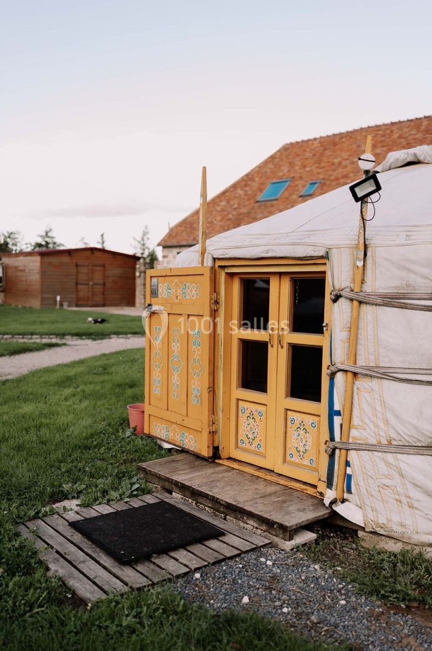 Yourte blanche avec porte en bois jaune ornée de motifs, située sur une pelouse près de bâtiments en arrière-plan.