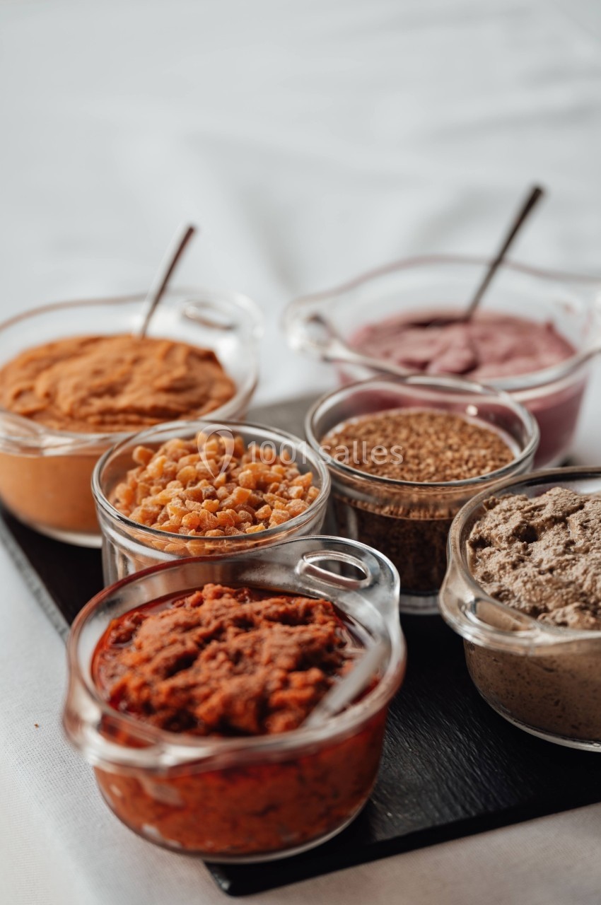 Assortiment de condiments et purées dans des bols en verre sur une planche noire, avec une nappe blanche en arrière-plan.